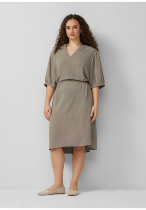 Kobieta o kręconych włosach, ubrana w luźną sukienkę w kolorze taupe sięgającą kolan z bufkami na rękawach i beżowe baleriny, stojąca na tle jednolitej szarej ściany.