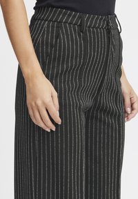 Črne prugaste hlače z metalno srebrnimi črtami, izdelane iz strukturirane tkanine. Imajo kroj, prilagojen postavi, in stranske žepe.