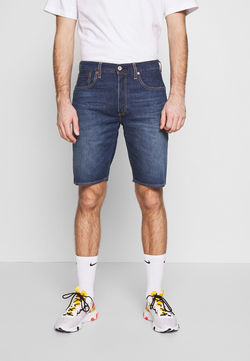 Homme portant un short en jean bleu foncé, des chaussettes blanches avec un logo noir, des baskets blanches avec des accents jaunes et oranges, et un t-shirt blanc.
