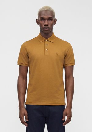 Homme aux cheveux blonds courts portant un polo marron moutarde et un pantalon bleu marine foncé, se tenant devant un fond clair uni.