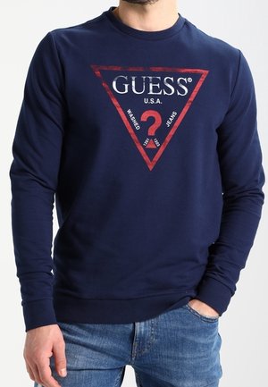 Mężczyzna w granatowej koszuli z długim rękawem z czerwonym trójkątnym logo z napisem "GUESS U.S.A. Washed Jeans" i niebieskich dżinsach.