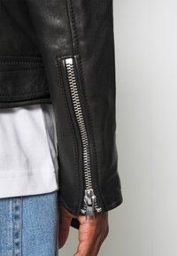 Sort læderjakkeærme med en sølvfarvet lynlåsdetalje. Glat texture og tætsiddende design. Hvid skjorte og blå denim er synlig nedenunder.