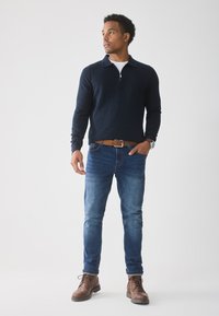 Man staat, draagt een navy kleurige ritsvest, blauwe jeans, een bruine riem, bruine laarzen en een horloge, kijkt opzij tegen een effen witte achtergrond.