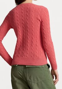 Różowy sweter w splot warkoczowy z prążkowanym dołem i mankietami, zestawiony z oliwkowozielonymi spodniami. Sweter ma teksturowany wzór i dopasowany krój.