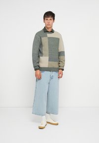 Maglione in varie tonalità di verde e crema con un motivo geometrico. Abbinato a jeans a gamba larga blu chiaro e sneakers bianche.