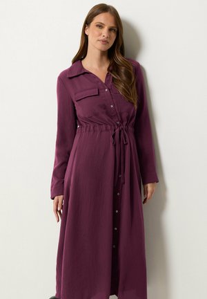 Femme aux longs cheveux bruns portant une robe violette à manches longues boutonnée avec une ceinture à la taille, debout contre un mur blanc.