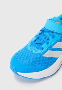 Zapatilla deportiva azul con parte superior de malla, superposiciones texturizadas y talón moldeado. Incluye acentos blancos contrastantes y correas ajustables.
