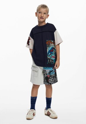 Navy plava i bijela majica kratkih rukava s grafičkim printom; svijetlosive cargo kratke hlače s stripovskim motivima; plave čarape; bijele tenisice s crvenim detaljima.