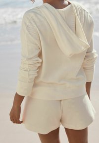 Una sudadera de punto waffle de color crema con capucha, combinada con pantalones cortos a juego. La tela es de textura suave y presenta mangas y puños enrollados.