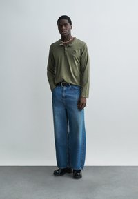 Férfi áll zsebébe dugott kézzel, olívazöld hosszú ujjú inget, széles szárú kék farmert, fekete cipőt és gyöngynyakláncot visel semleges háttér előtt.