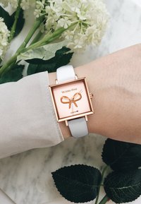 Reloj cuadrado con un marco de oro rosa, correa de cuero blanco y una esfera rosa con un diseño de lazo dorado. Contra un fondo de mármol.