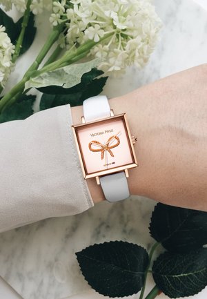 Orologio quadrato con cassa in oro rosa, cinturino in pelle bianca e quadrante rosa con un design a fiocco dorato. Posizionato su uno sfondo di marmo.