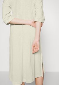 Personne portant une robe beige clair longueur genoux avec des manches trois-quarts, tenant son bras gauche derrière son dos.