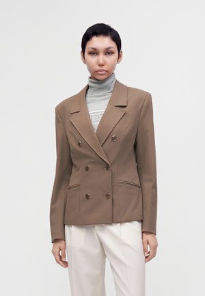 Persona joven con cabello corto y negro lleva una blazer marrón cruzada sobre un suéter de cuello alto gris y pantalones blancos, de pie frente a un fondo liso.