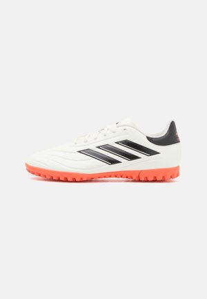Weißer und schwarzer flacher Fußballschuh mit oranger Sohle und strukturiertem Grip, von der Seite auf einfarbigem Hintergrund gesehen.