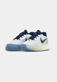 Αθλητικά παπούτσια Nike σε λευκό χρώμα με μπλε λεπτομέρειες, με υφασμάτινη επάνω επιφάνεια με υφή, μαύρο λογότυπο και ελαφρύ κίτρινο ενδιάμεσο σόλο με μοτίβο μπλε σόλας.