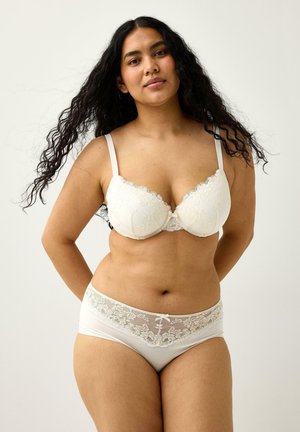 Kvinde med langt sort hår iført hvid blonde-bh og matchende underbukser med blonder, stående mod en ensfarvet baggrund.