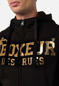 Felpa nera con zip realizzata in un mix di cotone, con stampa testurizzata dorata del logo "BOXEUR DES RUES" sul davanti e con lacci neri.