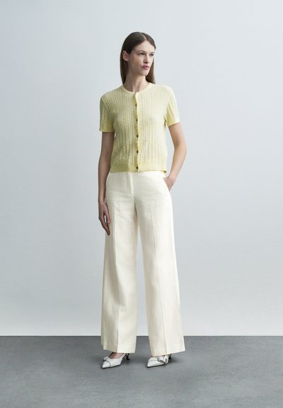 Γυναίκα που στέκεται φορώντας ένα ανοιχτό κίτρινο κοντομάνικο πλεκτό ζακέτα, κρεμ παντελόνα με φαρδιά μπατζάκια και λευκά παπούτσια με χαμηλό τακούνι, μπροστά σε απλό γκρι φόντο.