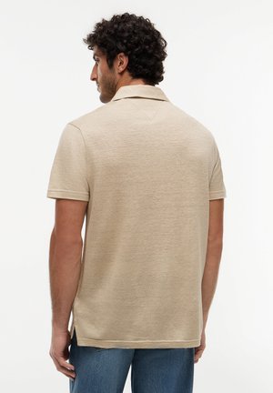 Homme aux cheveux bouclés et foncés, portant un polo beige texturé à manches courtes et un jean bleu, tourné dos à un fond uni.