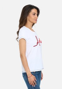 Camiseta de algodón blanca con cuello redondo, mangas cortas enrolladas y un diseño en rojo con escritura en la parte frontal. Llevada con jeans de mezclilla azules.