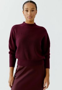 Burgunderfarbener Strickpullover mit hohem Kragen, geripptem Muster und überschnittenen Schultern, kombiniert mit einem passenden, figurbetonten Rock.