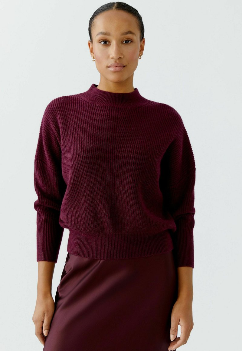 Burgunderfarbener Strickpullover mit hohem Kragen, geripptem Muster und überschnittenen Schultern, kombiniert mit einem passenden, figurbetonten Rock.