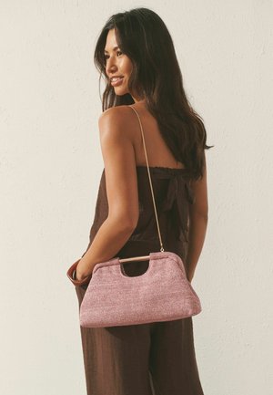 Woman wearing brown strapless outfit holding pink textured handbag with gold chain strap, standing against a light textured wall.