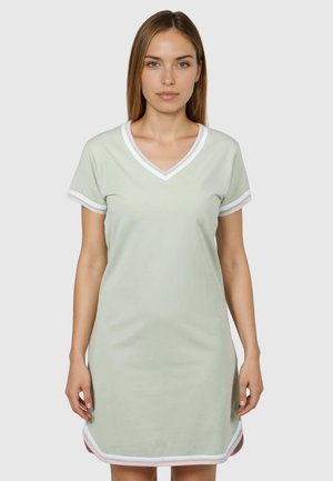 Kvinde iført lysegrøn kortærmet V-hals kjole med hvid kant, stående foran ensfarvet baggrund.