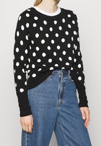 Black sweater with white polka dots, featuring a crew neck, long sleeves, and ribbed cuffs, paired with high-waisted blue jeans.