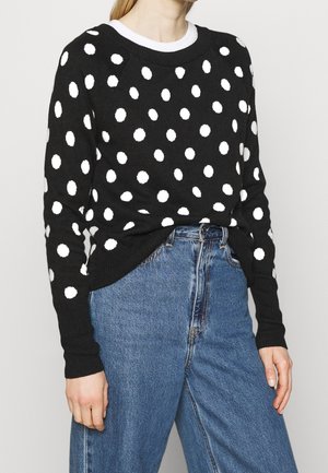Pull noir avec des pois blancs, doté d'un col rond, de manches longues et de poignets côtelés, associé à un jean bleu taille haute.