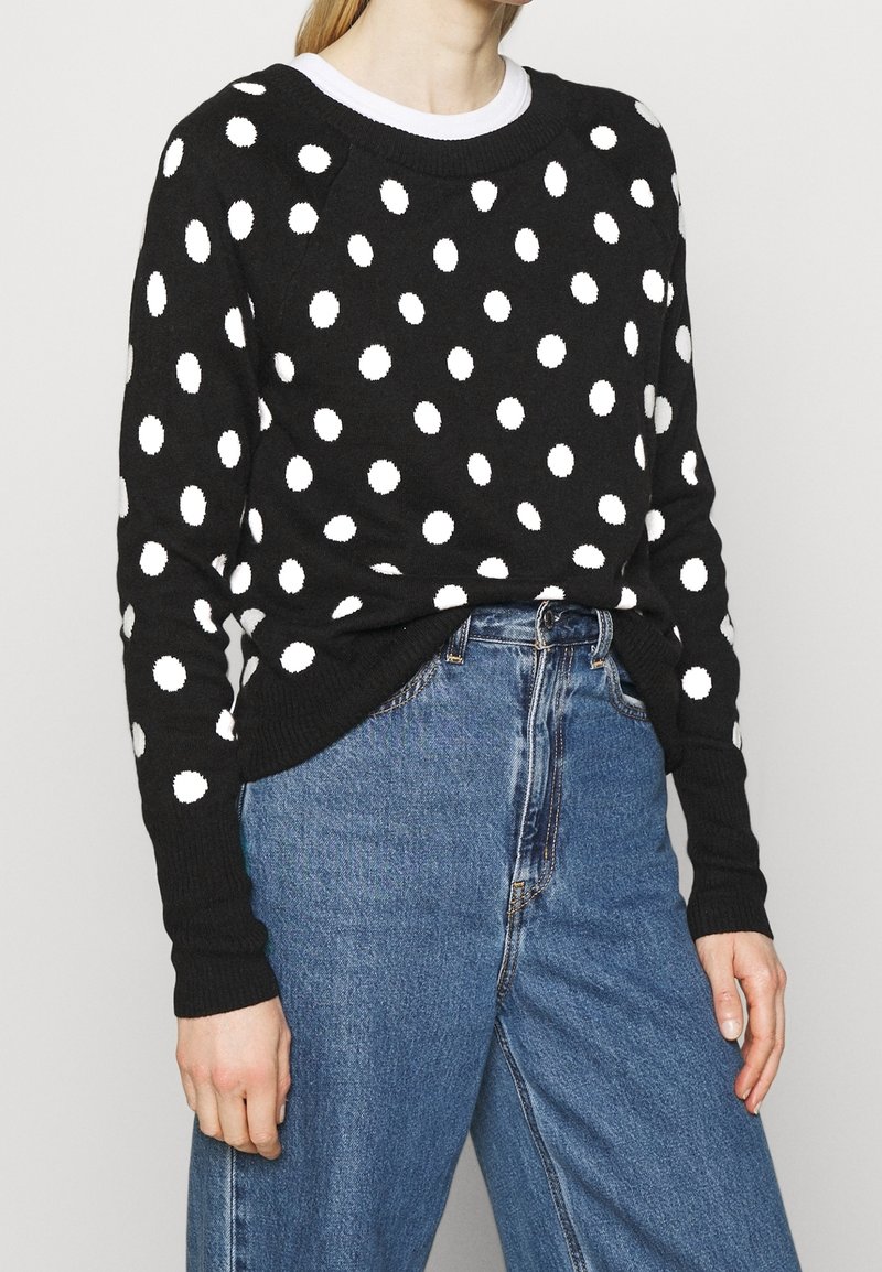Black sweater with white polka dots, featuring a crew neck, long sleeves, and ribbed cuffs, paired with high-waisted blue jeans.