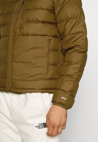 Brown puffer jacket with a zippered chest pocket, ribbed cuffs, and a "550" detail on the sleeve. Worn with white fleece pants.