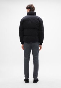 Jachetă puf din negru cu guler înalt, secțiuni matlasate și manșete elastice, purtată peste pantaloni gri și pantofi negri. Fundal simplu.