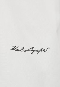 Μαύρη υπογραφή με καλλιγραφικά γράμματα "Karl Lagerfeld" στο κέντρο σε απλό λευκό φόντο, πιθανόν ετικέτα υφάσματος ή ρούχου.