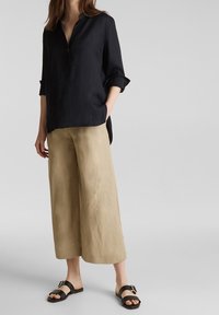 Frau in einem lockeren schwarzen Langarmshirt, hellbeigen weit geschnittenen Caprihosen und schwarzen Rutschschuhsandalen mit Schnallen, die auf grauem Boden steht.