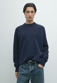 Junger Mann, der einen marineblauen Pullover und blaue Jeans trägt, steht mit einer Hand in der Tasche vor einem schlichten hellen Hintergrund.