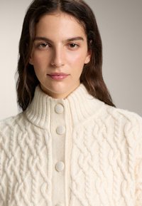 Krem pulover s pletenim vzorcem iz vrvi, visokim ovratnikom in tremi velikimi, teksturiranimi gumbi. Mehka volna z zapletenimi upognjenimi vzorci.