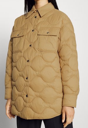 Lysebrun quiltet jakke med bølget syning, knaplukning foran, to brystlommer med klapper og knapper, båret over sorte bukser.