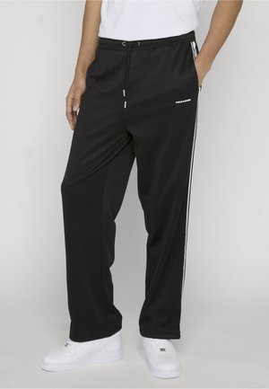 Sorte træningsbukser med hvide sidelister og sidelommer, båret med hvide sneakers og en hvid skjorte, som er stukket ned.