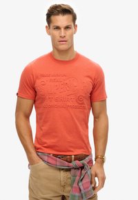 Short-sleeve, coral red T-shirt with embossed brand text, paired with khaki pants. A plaid shirt is tied around the waist.