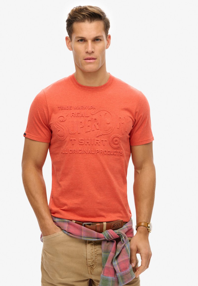 Short-sleeve, coral red T-shirt with embossed brand text, paired with khaki pants. A plaid shirt is tied around the waist.