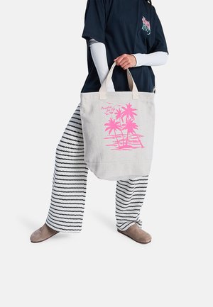 Persona con camiseta negra y pantalones a rayas sostiene una bolsa tote beige con diseño de palmera rosa y texto "paraíso para siempre."