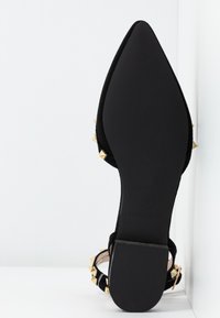 Chaussure plate noire pointue avec bride à la cheville, décorée de clous pyramides dorés, semelle visible, posée sur fond blanc.