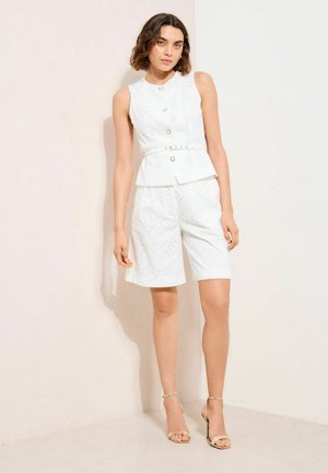 Femeie care stă lângă un perete deschis la culoare, purtând un top alb fără mâneci, cu nasturi și curea, și pantaloni scurți până la genunchi asortați, cu sandale cu toc înalt.