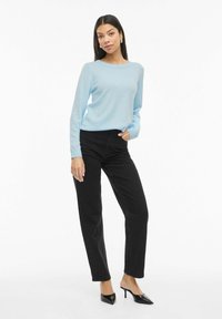 Femme portant un pull bleu clair, un jean noir taille haute et des mules à talons noires, debout avec une main dans la poche sur fond blanc.