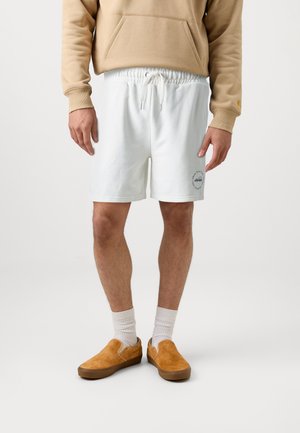 Pantalones cortos de algodón blancos con cintura ajustable y un pequeño detalle de logo. Combinados con zapatos de ante color beige sin cordones y calcetines blancos de canalé.
