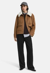 Braune Wolljacke mit weißem Kunstfellkragen, ausgestattet mit zwei vorderen Taschen und einem Reißverschluss. Kombiniert mit einem dunkelbraunen Pullover und lockeren schwarzen Hosen.