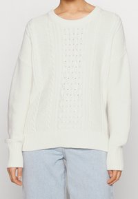 Witte gebreide trui met een mix van kabel- en structuurpatronen, een ronde hals en geribbelde boorden en zoom. Gecombineerd met lichtblauwe spijkerbroeken.