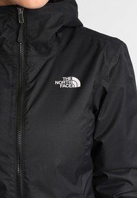 Svart ytterjacka i slitstarkt material med dragkedja och logo på bröstet. Den har huva och slät textur.